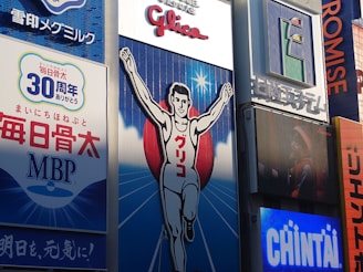 A large billboard featuring an illustration of a runner with arms raised in victory, set against a blue background with white rays emanating from the center. The surrounding area includes various advertisements and signs containing Japanese text, some with corporate logos.