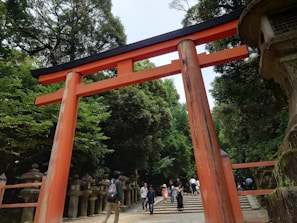 A large red torii gate stands prominently in the foreground, located in a lush, green forest setting. Several stone lanterns line the pathway leading to stairs in the background, where people are walking and exploring the area. The atmosphere is serene, combining natural beauty with historical architecture.