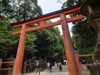 A large red torii gate stands prominently in the foreground, located in a lush, green forest setting. Several stone lanterns line the pathway leading to stairs in the background, where people are walking and exploring the area. The atmosphere is serene, combining natural beauty with historical architecture.