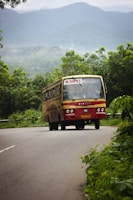 A red and cream KSRTC bus is traveling down a winding rural road surrounded by lush green vegetation. The mountainous backdrop is shrouded in mist, suggesting a serene and natural setting. The road curves gently, contributing to the scenic and peaceful vibe.