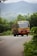 A red and cream KSRTC bus is traveling down a winding rural road surrounded by lush green vegetation. The mountainous backdrop is shrouded in mist, suggesting a serene and natural setting. The road curves gently, contributing to the scenic and peaceful vibe.