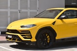 A yellow luxury SUV is parked near a white shuttered building. The car features sleek, modern design elements with black accents on the wheels and a glossy finish.
