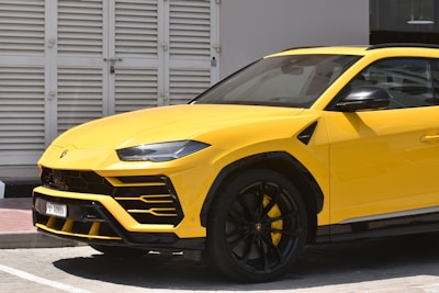 A yellow luxury SUV is parked near a white shuttered building. The car features sleek, modern design elements with black accents on the wheels and a glossy finish.