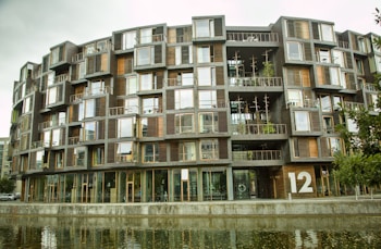 A modern apartment building with a unique structural design featuring numerous windows and balconies. It has a natural wood and glass facade, and several plants are visible on the balconies. The building is adjacent to a water body, reflecting its structure.