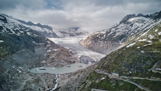Snow-capped mountains surround a wide glacier that flows into a rocky valley below. Patches of green vegetation are visible on the slopes, and a narrow, winding road leads to a small structure near the foreground. The sky is overcast, creating a dramatic contrast with the rugged landscape.