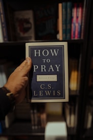 A hand holding a book titled 'How to Pray' by C.S. Lewis, displayed against a backdrop of books on a shelf.