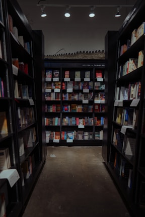 A well-organized aisle in a bookstore with shelves lined with a variety of books. The shelves are filled with colorful book covers, and there's a section labeled 'Mystery/Thriller'. Overhead lighting casts a warm glow on the book displays.