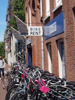A row of bicycles is lined up against a brick building with signs indicating a bike rental shop. The shop signs are visible in different languages, and the brick structure suggests a traditional or historic urban setting. A person with a backpack walks nearby on the sidewalk. The atmosphere suggests a casual and leisurely urban environment.