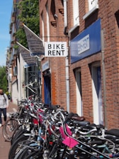 A row of bicycles is lined up against a brick building with signs indicating a bike rental shop. The shop signs are visible in different languages, and the brick structure suggests a traditional or historic urban setting. A person with a backpack walks nearby on the sidewalk. The atmosphere suggests a casual and leisurely urban environment.
