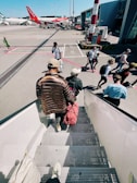 Passengers are seen descending the stairs from an aircraft and walking across the tarmac towards an airport terminal. Several people carry backpacks and travel bags. In the background, there are airplanes parked, with one red aircraft marked with the easyJet logo. The scene captures a typical boarding or disembarking scenario at an airport on a clear day.