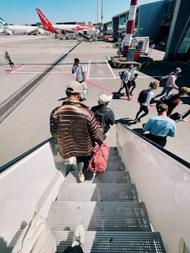 Passengers are seen descending the stairs from an aircraft and walking across the tarmac towards an airport terminal. Several people carry backpacks and travel bags. In the background, there are airplanes parked, with one red aircraft marked with the easyJet logo. The scene captures a typical boarding or disembarking scenario at an airport on a clear day.