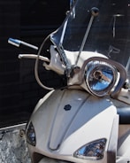 A close-up view of the front of a motor scooter, featuring a clear windshield, headlight, and handlebars. The scooter is white with a logo on the front panel, and its surface shows some signs of dirt and wear.