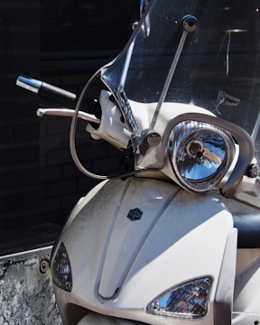 A close-up view of the front of a motor scooter, featuring a clear windshield, headlight, and handlebars. The scooter is white with a logo on the front panel, and its surface shows some signs of dirt and wear.
