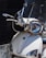 A close-up view of the front of a motor scooter, featuring a clear windshield, headlight, and handlebars. The scooter is white with a logo on the front panel, and its surface shows some signs of dirt and wear.