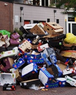 A large pile of discarded cardboard boxes, many labeled with fruit brands, stacked on a street corner near a brick building. The boxes are various colors, and some yellow plastic bags are mixed in. The background includes a modern building facade with windows and some greenery.