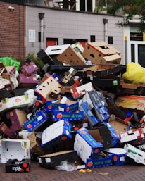 A large pile of discarded cardboard boxes, many labeled with fruit brands, stacked on a street corner near a brick building. The boxes are various colors, and some yellow plastic bags are mixed in. The background includes a modern building facade with windows and some greenery.