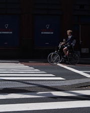 A person rides a bicycle across a street while a pedestrian crosswalk is visible in the foreground. The background features a dark building facade with signage. The lighting casts shadows on the road, creating a contrast between the bright crosswalk and the darker surroundings.