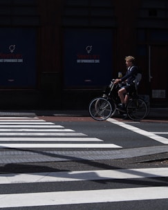 A person rides a bicycle across a street while a pedestrian crosswalk is visible in the foreground. The background features a dark building facade with signage. The lighting casts shadows on the road, creating a contrast between the bright crosswalk and the darker surroundings.