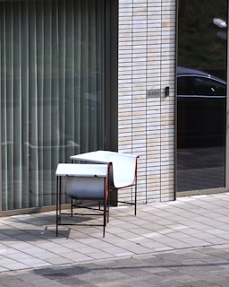 A white chair with a red frame and a small metal table are placed on a tiled patio. The setting is in front of a modern building with large glass windows and vertical blinds, alongside a brick wall with a label indicating '44B1'. A parked car is reflected in the glass.