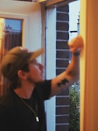 A person with a cap is reaching towards a door or window frame, possibly cleaning or inspecting it. The setting appears to be indoors but with a clear view of an outside scene featuring greenery and a brick wall.