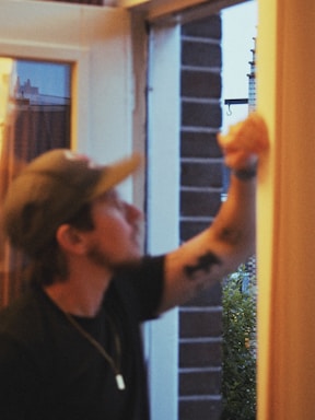 A person with a cap is reaching towards a door or window frame, possibly cleaning or inspecting it. The setting appears to be indoors but with a clear view of an outside scene featuring greenery and a brick wall.