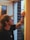 A person with a cap is reaching towards a door or window frame, possibly cleaning or inspecting it. The setting appears to be indoors but with a clear view of an outside scene featuring greenery and a brick wall.