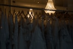 A collection of elegant wedding dresses hanging on wooden hangers is displayed in a dimly lit room. The dresses feature intricate lace patterns and delicate fabric. Above them, a luxurious crystal chandelier casts a warm, soft glow, adding a touch of sophistication and elegance to the scene.
