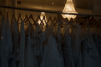 A collection of elegant wedding dresses hanging on wooden hangers is displayed in a dimly lit room. The dresses feature intricate lace patterns and delicate fabric. Above them, a luxurious crystal chandelier casts a warm, soft glow, adding a touch of sophistication and elegance to the scene.