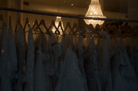 A collection of elegant wedding dresses hanging on wooden hangers is displayed in a dimly lit room. The dresses feature intricate lace patterns and delicate fabric. Above them, a luxurious crystal chandelier casts a warm, soft glow, adding a touch of sophistication and elegance to the scene.