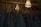 A collection of elegant wedding dresses hanging on wooden hangers is displayed in a dimly lit room. The dresses feature intricate lace patterns and delicate fabric. Above them, a luxurious crystal chandelier casts a warm, soft glow, adding a touch of sophistication and elegance to the scene.