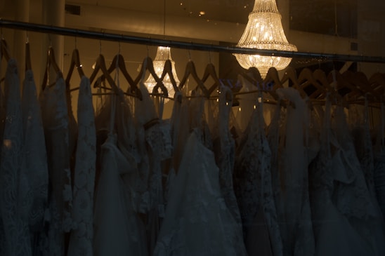 A collection of elegant wedding dresses hanging on wooden hangers is displayed in a dimly lit room. The dresses feature intricate lace patterns and delicate fabric. Above them, a luxurious crystal chandelier casts a warm, soft glow, adding a touch of sophistication and elegance to the scene.