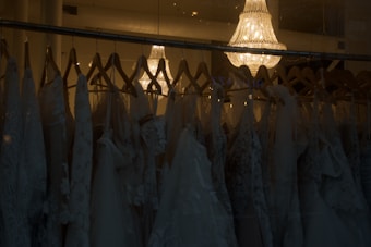 A collection of elegant wedding dresses hanging on wooden hangers is displayed in a dimly lit room. The dresses feature intricate lace patterns and delicate fabric. Above them, a luxurious crystal chandelier casts a warm, soft glow, adding a touch of sophistication and elegance to the scene.