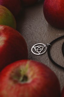 Several red apples are placed around a silver pendant with intricate circular patterns, lying on a textured surface. The pendant is attached to a black cord.