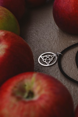 Several red apples are placed around a silver pendant with intricate circular patterns, lying on a textured surface. The pendant is attached to a black cord.