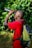 A person wearing a red shirt and a backpack is looking through binoculars. The background consists of lush green foliage, suggesting a natural outdoor setting.