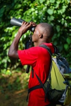 A person wearing a red shirt and a backpack is looking through binoculars. The background consists of lush green foliage, suggesting a natural outdoor setting.