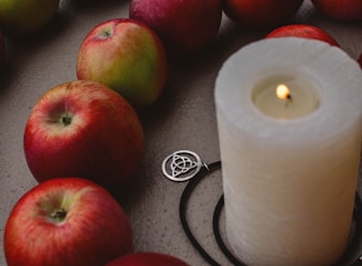 A group of red apples arranged around a large lit white candle, which is accompanied by a silver triquetra pendant. The scene is set against a textured, neutral-colored surface.