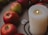 A group of red apples arranged around a large lit white candle, which is accompanied by a silver triquetra pendant. The scene is set against a textured, neutral-colored surface.