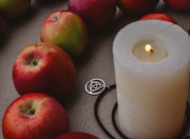 A group of red apples arranged around a large lit white candle, which is accompanied by a silver triquetra pendant. The scene is set against a textured, neutral-colored surface.