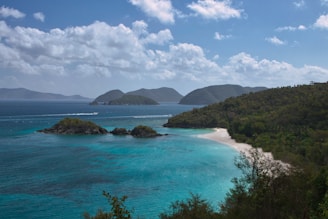 A serene tropical beach with lush greenery, a white sandy shoreline, and turquoise blue waters. Small islands are visible in the distance under a bright blue sky with scattered fluffy clouds.