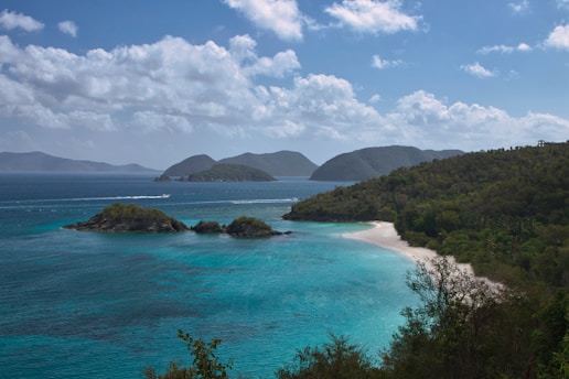 A serene tropical beach with lush greenery, a white sandy shoreline, and turquoise blue waters. Small islands are visible in the distance under a bright blue sky with scattered fluffy clouds.