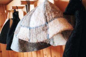 Several wool hats hang on wooden pegs against a wooden background, with a focus on a hat with a plaid pattern in shades of gray, cream, and blue.