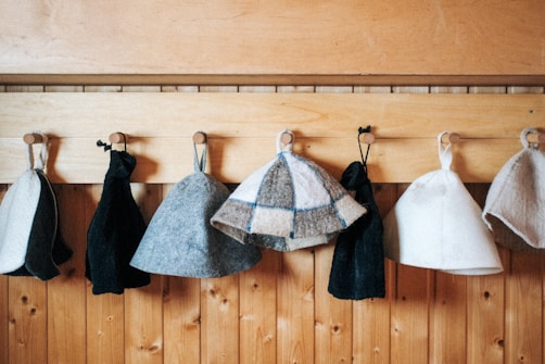 A series of woolen hats and black pouches hang on wooden pegs attached to a vertically paneled wooden wall. The hats vary in color including grey, white, and a gray checkered pattern, while the pouches are black.