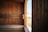 A room with wooden paneling and benches, typically used for a sauna. The wood has a warm, natural finish and there is natural light coming through a window on the right side. The window provides a view of an outdoor scene with some buildings and greenery in the background.