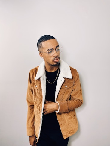 A person stands against a plain background wearing a brown shearling jacket over a black shirt. They have short hair and are wearing glasses. The person appears to be looking to the side with a thoughtful expression.