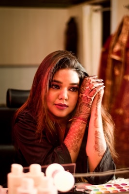 A woman with intricate henna designs on her hands is sitting and leaning her arms on a surface. She is wearing black attire, and her long hair is styled loosely. In the background, there are blurred objects including pieces of clothing. The foreground shows several small bottles and a palette with various colors, indicating a makeup or dressing room setting.
