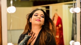 A person with long hair is in front of a mirror, seemingly styled for an occasion with a bright makeup look. They are holding a hair dryer and wearing a black outfit. The background shows a blurred garment, which is red with gold details, hanging on the wall.