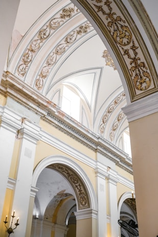 Ornate architectural arches adorned with intricate gold and tan designs on a cream and white background. The structure features grand columns and high vaulted ceilings, with elegant chandeliers barely visible in the lower corners.