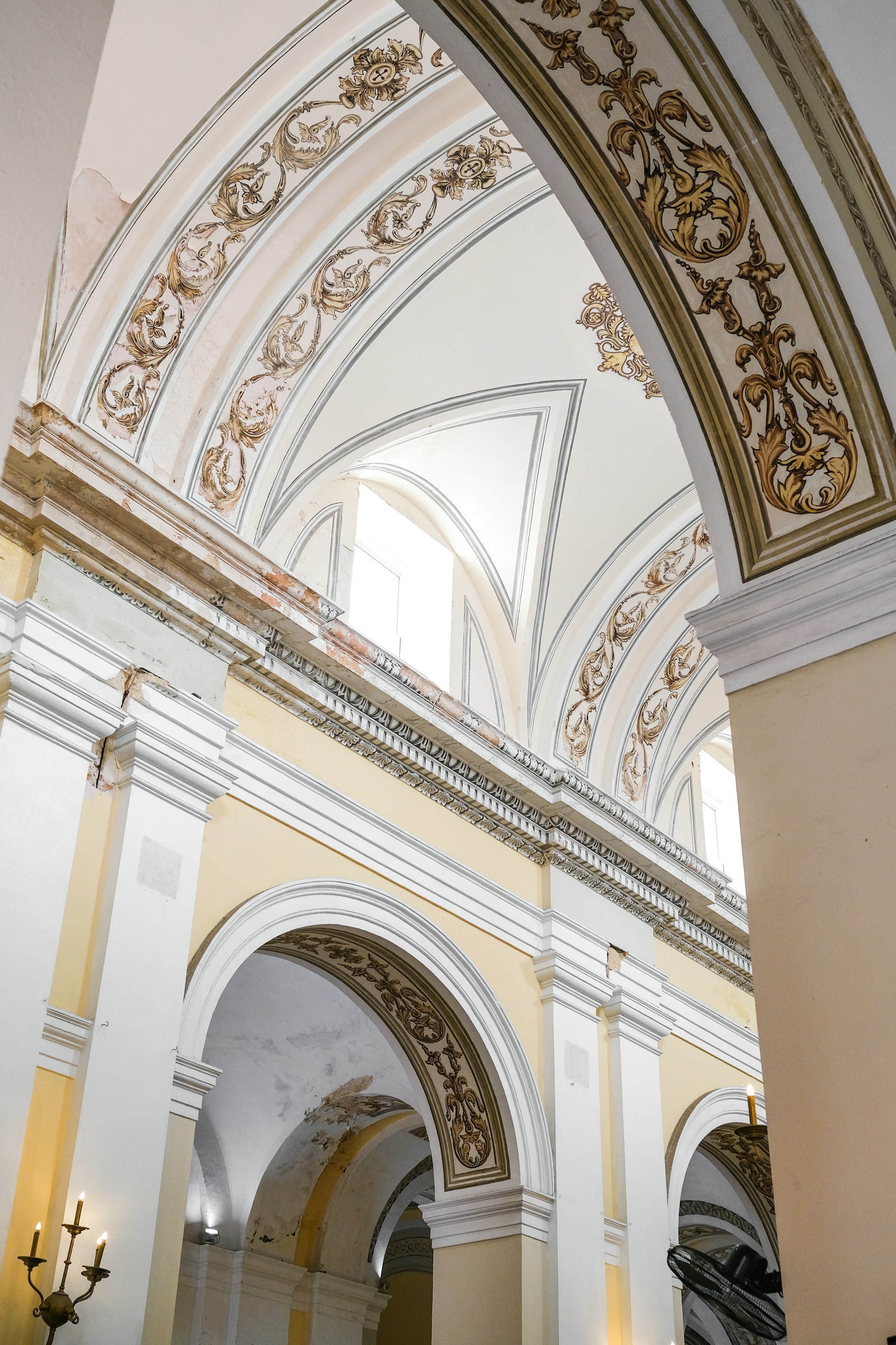 Ornate architectural arches adorned with intricate gold and tan designs on a cream and white background. The structure features grand columns and high vaulted ceilings, with elegant chandeliers barely visible in the lower corners.