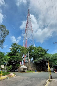 A tall red and white communication tower rises high into a partly cloudy blue sky. The base of the tower is surrounded by trees and a small white tent. A couple of parked cars are visible near the tent, and some road signs and barriers are present at the intersection in the foreground.